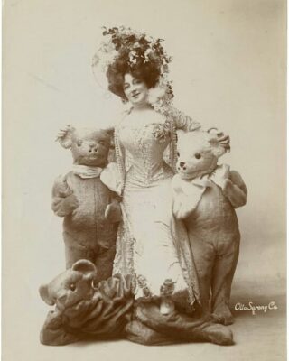 En viktoriansk kvinde med en kæmpe hat med blomster omgivet af 3 børn i bamsedragter med lille talje og blød hud.